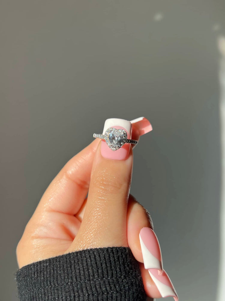 A woman’s hand with French manicure nails holds a silver ring featuring a large heart-shaped diamond. The ring is adorned with small diamonds along the band. Her hand is dressed in a black sweater, creating a striking contrast with the ring's brilliance