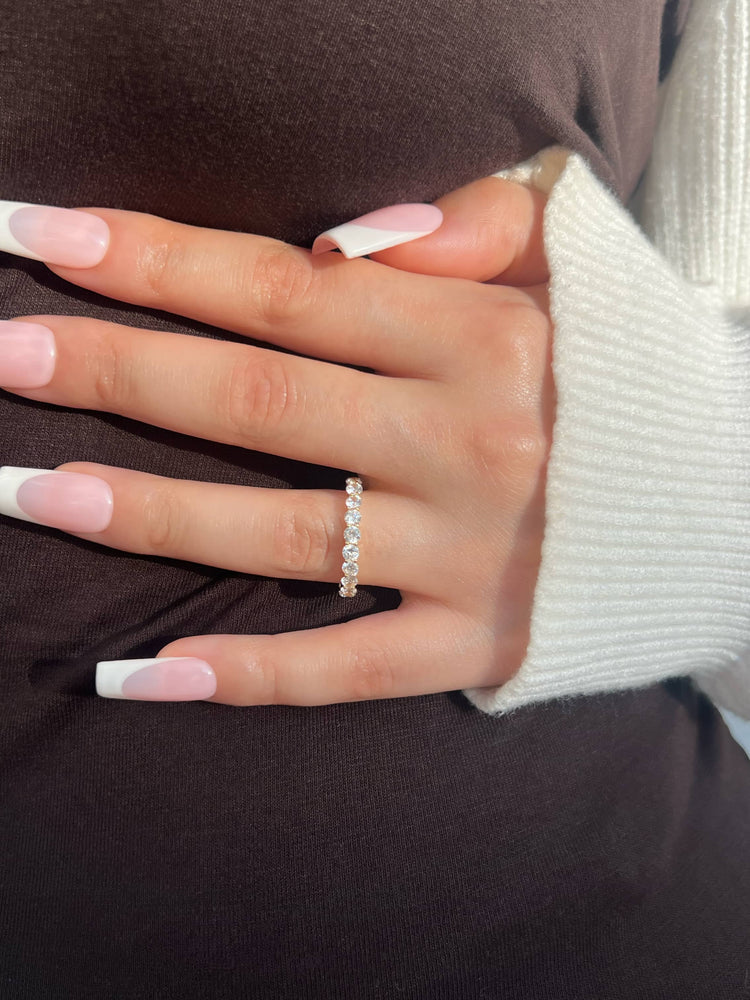 A woman’s hand with long fresh manicured nails wearing a 3mm thin gold band with clear round stones. The hand rests on a dark brown dress, with a white sweater layered on her hand, showcasing an elegant and stylish look.