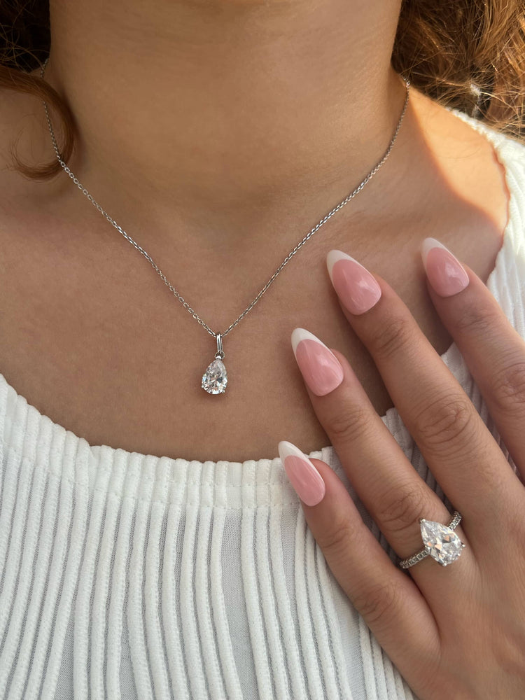 A woman wearing a white top shows off her left hand, featuring a silver ring with a pear-shaped diamond. Her nails are polished with a French manicure. She also wears a silver necklace with a matching pear-shaped diamond pendant, for an elegant look.