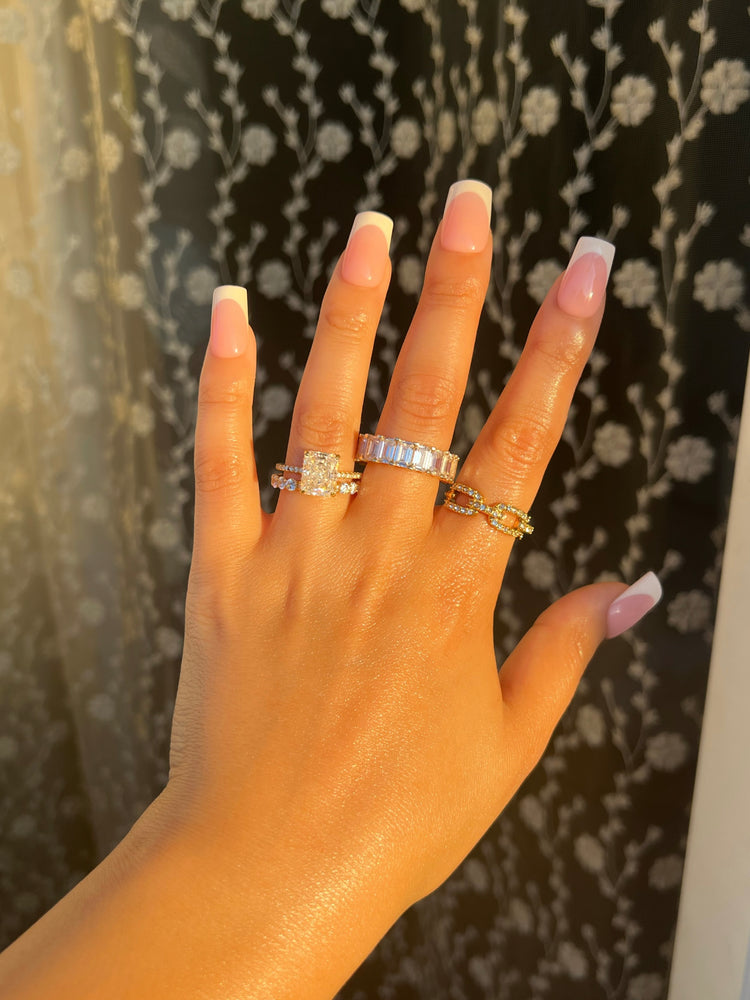 A woman’s hand with white French manicured nails showcases four rings: a large radiant cut shaped diamond ring, a 3mm diamond eternity gold band, an emerald cut band with rectangular diamonds, and a chain link ring, creating a luxury, elegant ring stack.