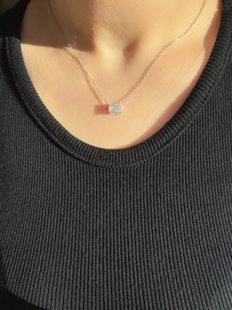 A woman wearing a black ribbed top adorned with a silver chain necklace. The necklace features a large radiant cut rectangular clear stone pendant that catches the light beautifully, adding a touch of elegance to the outfit