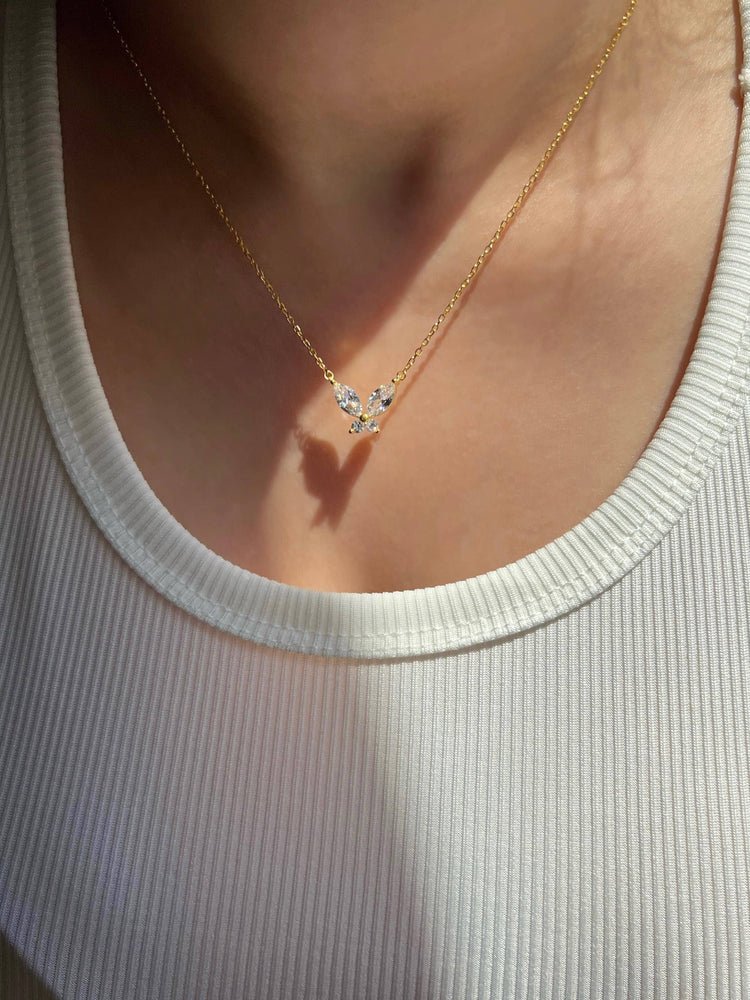 A woman is wearing a white ribbed tank top adorned with a gold chain necklace featuring a diamond butterfly pendant. The necklace casts a shadow on the surface below, creating a butterfly shape, adding a whimsical touch to the elegant design.