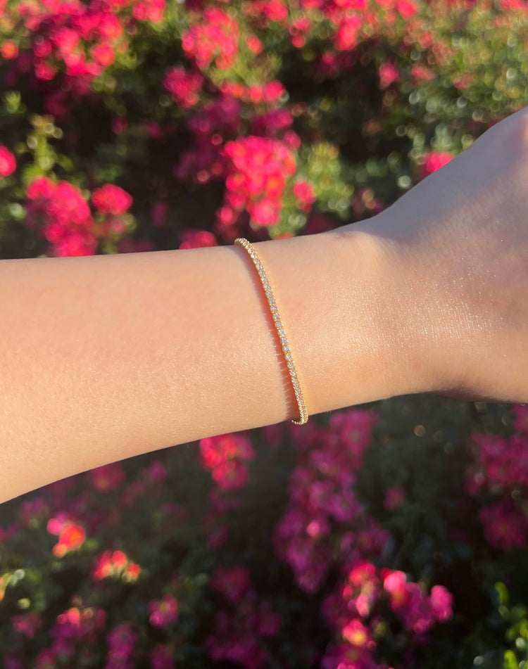 A close-up of a woman’s arm wearing a gold 2mm bracelet with small, clear stones encrusted along the entire band. The background features a soft blur of pink flowers, enhancing the bracelet's elegance and sparkle.