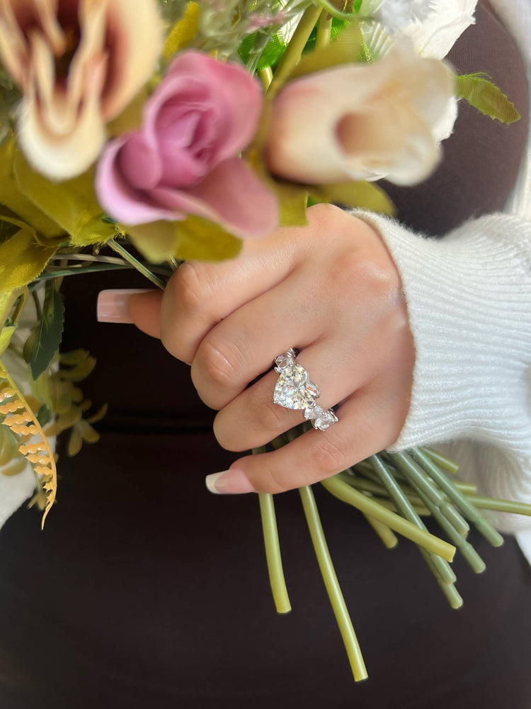 A woman’s hand in a white sweater holding a bouquet of pink and white roses, with green stems. The focus is on a large heart-shaped diamond ring on the ring finger, featuring one centre large heart-shaped diamond and smaller heart diamonds around the band