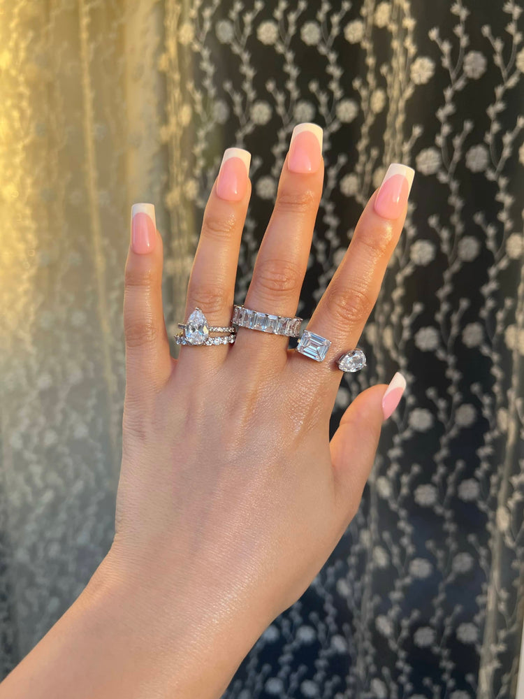 A woman’s hand adorned with four diamond rings; a pear-shaped diamond ring with a band of paved diamonds, a 4*6mm rectangular eternity band, a toi et moi rectangular and pear-shaped diamond ring. She has a short French manicure. Soft, neutral, background