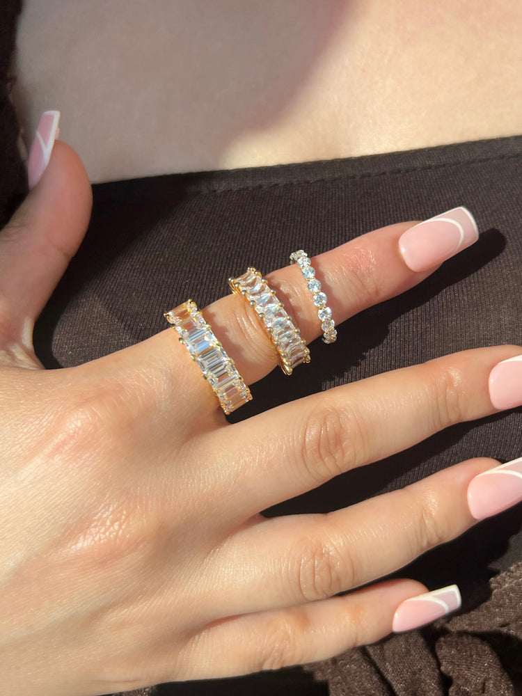 A close-up of a woman’s hand adorned with three gold clear stone rings. One ring showcases elegant round stones, while the other two rings are embellished with sparkling rectangular stones, creating a beautiful and eye-catching ring display.