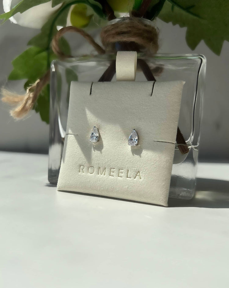 Elegant silver teardrop-shaped diamond earrings displayed on a white card labeled "ROMEELA." The earrings are showcased against a backdrop of a glass vase featuring a white flower and green leaves, highlighting their beauty and sophistication. 