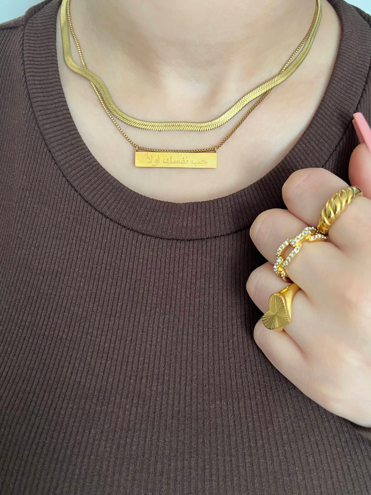 Woman in a brown ribbed top wearing a gold snake chain necklace and a rectangular pendant with the Arabic text ‘حب نفسك اولاً translating to ‘love yourself first’, and three gold rings on her right hand; a heart burst ring, link pave ring, croissant ring