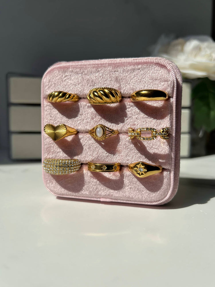 A collection of nine 18K Gold plated stainless steel rings displayed on a pink velvet tray. The rings vary in style, some are signet, paved, croissant, band and swirl design. They are arranged with three rings on each row, showcasing their unique designs