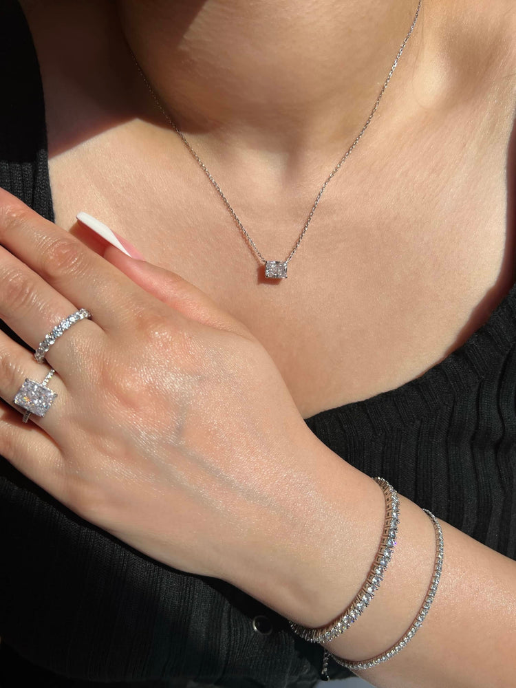 A woman wearing a black top with a rectangle diamond pendant silver chain necklace, two silver rings; a radiant cut diamond and a round cut eternity band, and two silver diamond tennis bracelets on her wrist. An elegant and sophisticated jewellery look