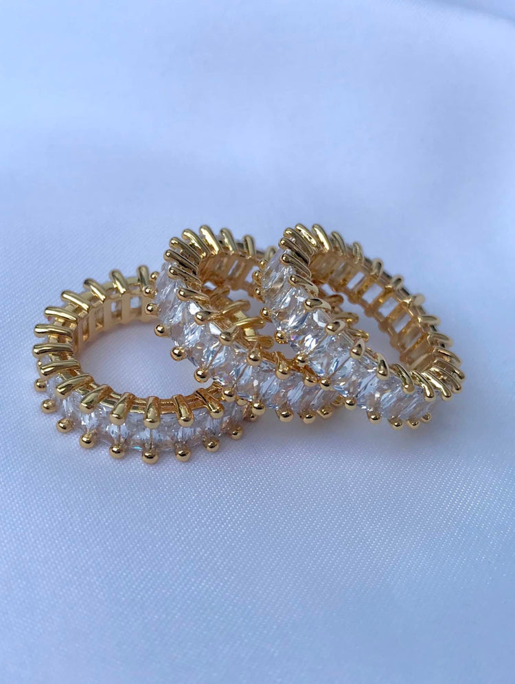 A close-up of three elegant gold rings featuring rows of clear, baguette-shaped cubic zirconia stones. The rings are arranged on a textured white surface, with the closest ring prominently displayed, showcasing its luxurious appearance and sparkly design