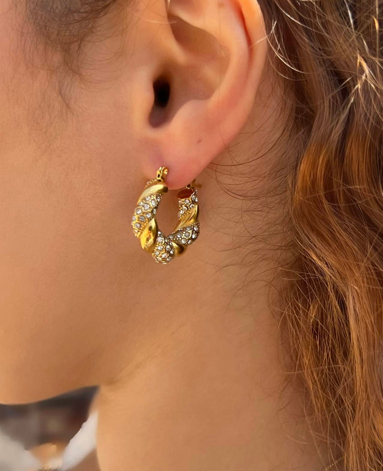 Woman wearing gorgeous gold hoop earring featuring a twisted design adorned with clear rhinestones. The earring's intricate detailing and sparkle enhance its elegance, making it a perfect accessory for any outfit or occasion.