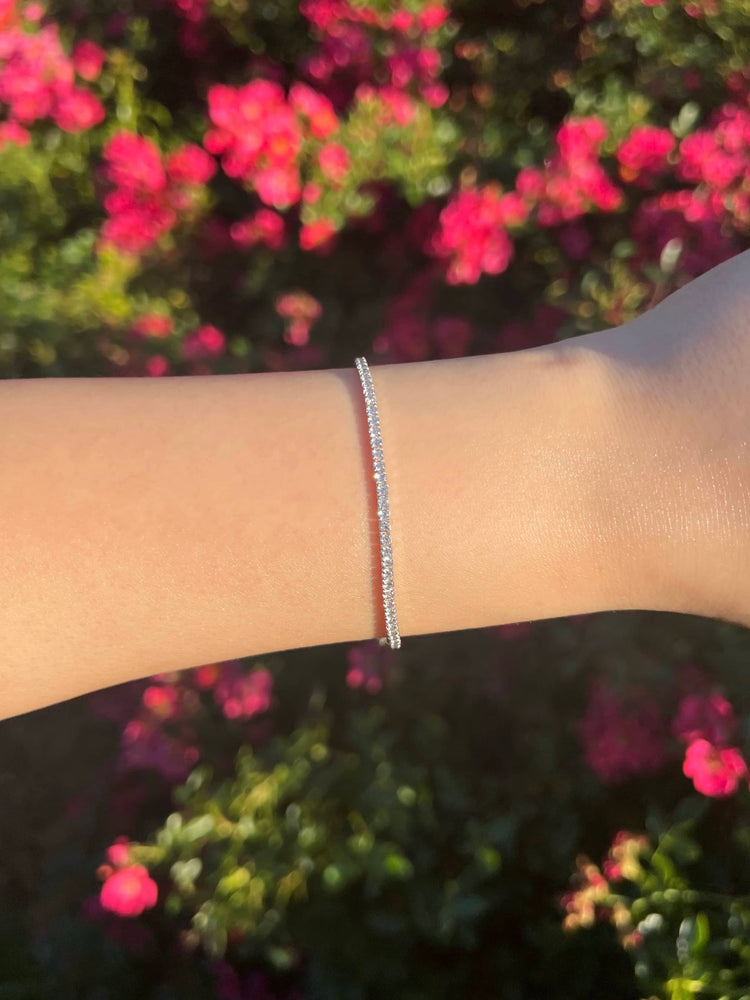 A woman's arm adorned with a thin 2mm silver bracelet featuring small diamonds. The background is softly blurred with pink flowers, adding a touch of elegance and color to the scene