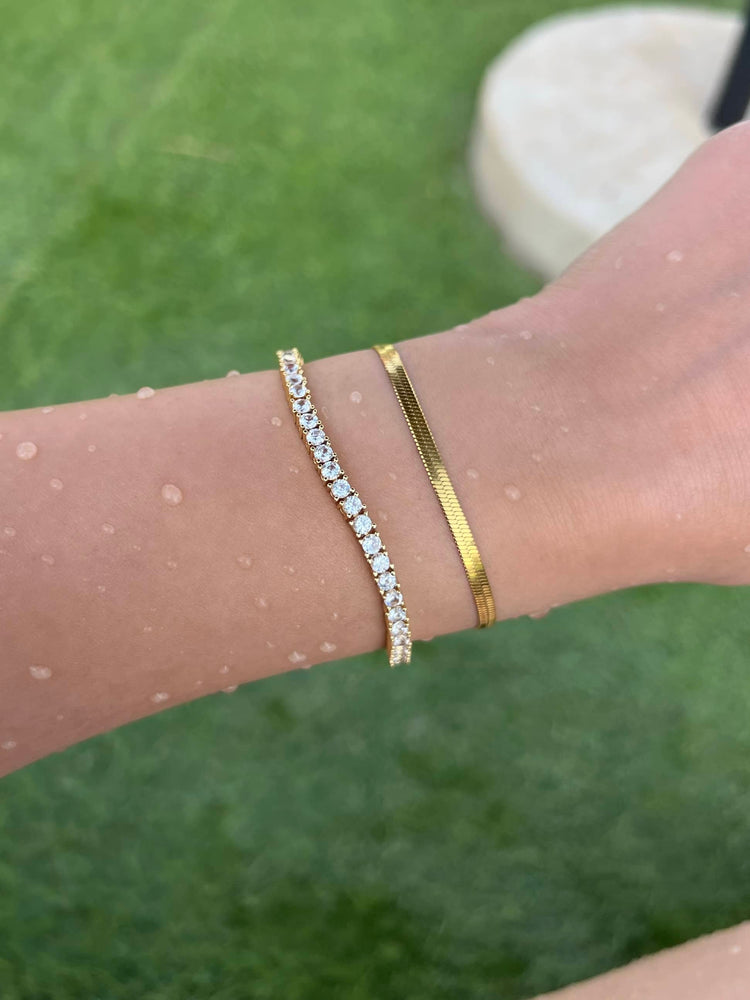 A woman's arm adorned with two gold bracelets. The first bracelet is a thin gold chain bracelet with a snake herringbone design, and the second is a 3mm tennis chain bracelet. Water droplets add a touch of elegance and show the bracelets are waterproof.  