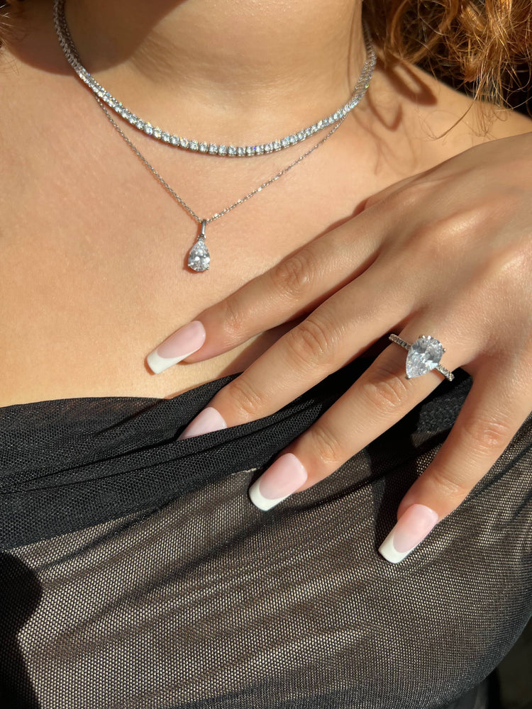 Woman in a black sheer top, wearing a silver tennis necklace, silver teardrop pendant necklace, silver pear-shaped diamond ring on ring finger, French manicured nails, showcasing a luxurious, elegant, date-night, sparkly look. 