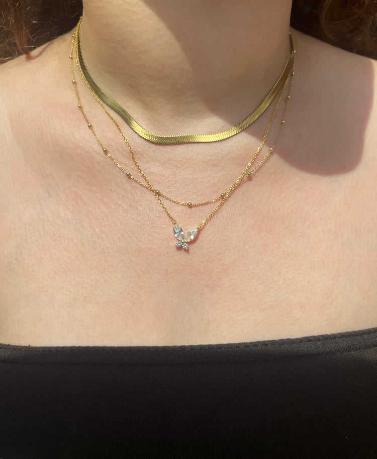 A woman wearing a dark brown bandeau top adorned with three gold necklaces. The first is a delicate snake chain, the second is a chain with small round beads, and the third features a charming butterfly pendant, creating a layered and fashionable look.