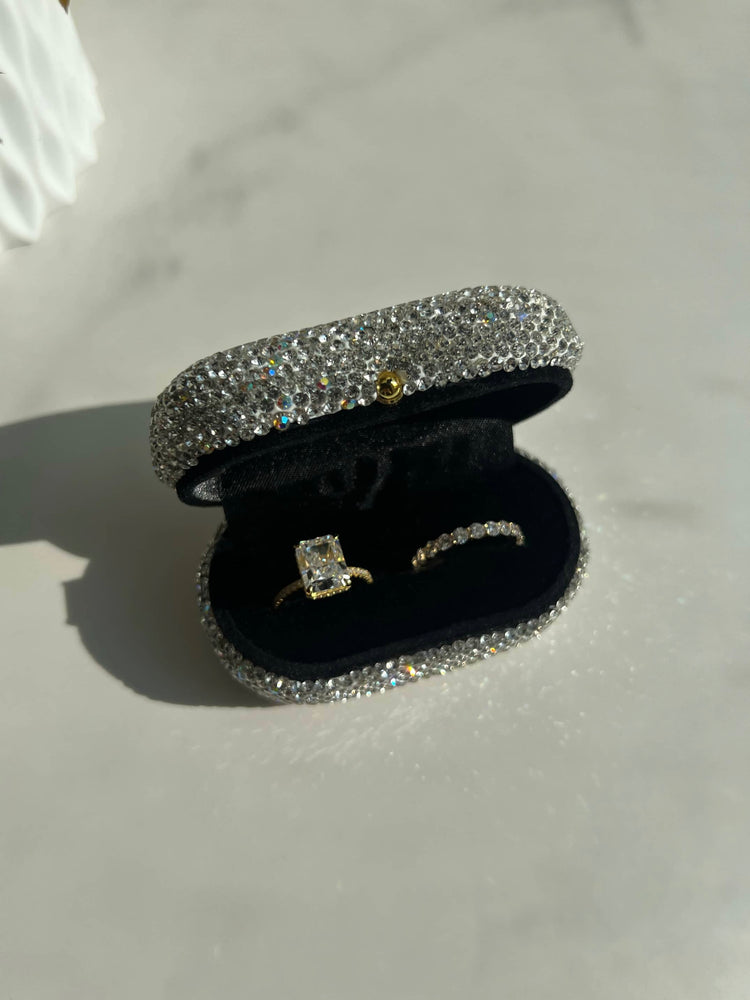 An elegant ring box shaped like an oval, covered in sparkling rhinestones, showcasing a stunning gold radiant diamond ring and a gold round cut wedding band inside, perfect for a special occasion or proposal