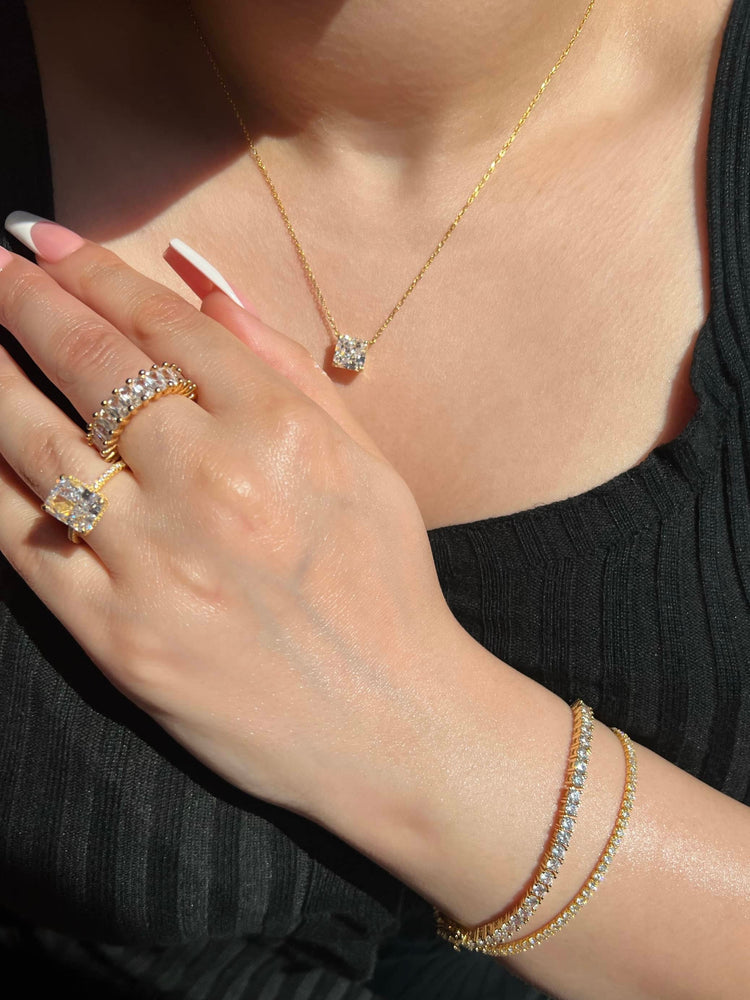 A woman wearing two gold diamond rings on her hand, one radiant cut and one eternity ring. She is also wearing a diamond pendant gold necklace, and two gold diamond tennis bracelets on her wrist. She is wearing a black top, showcasing luxury & elegance.