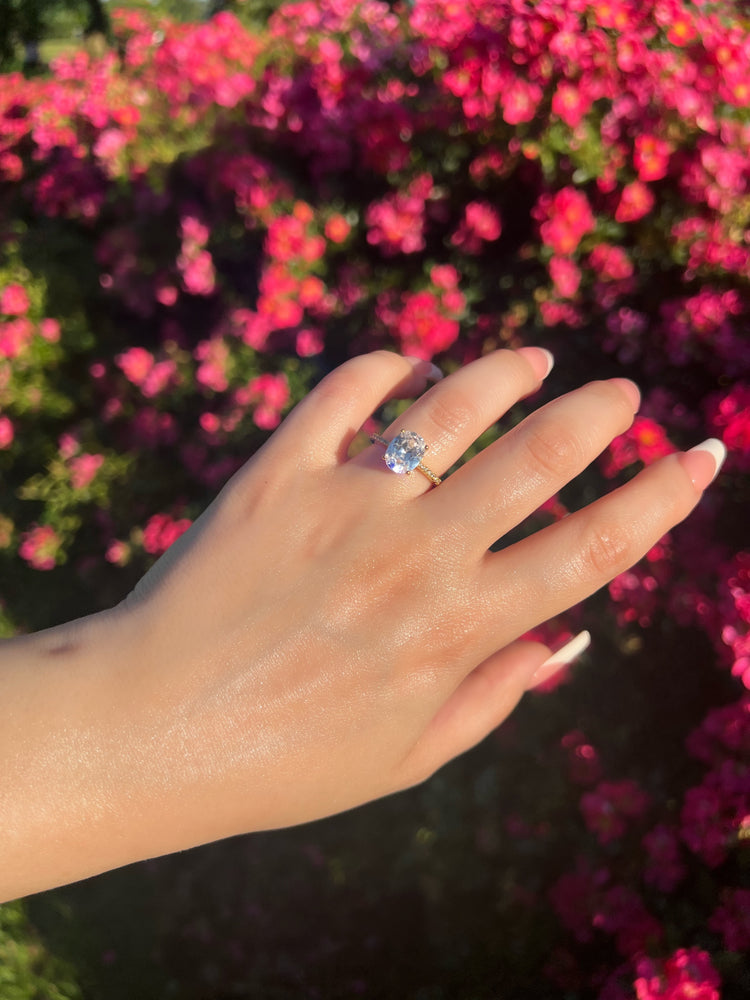 A close-up of a woman’s hand wearing a beautiful 2CT gold oval-shaped diamond ring. The ring features a thin band paved with small diamonds surrounding the large oval diamond, set against a soft, blurred background of pink flowers, enhancing its elegance.